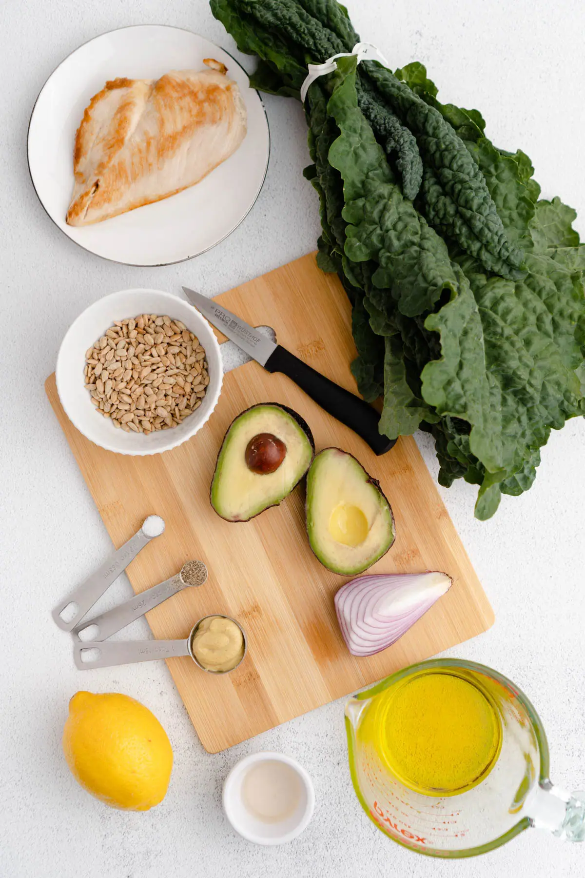 Overhead shot of ingredients used to prepare recipe: kale bunch, chicken breast on plate, olive oil in cup, lemon, cut avocado and red onion. 
