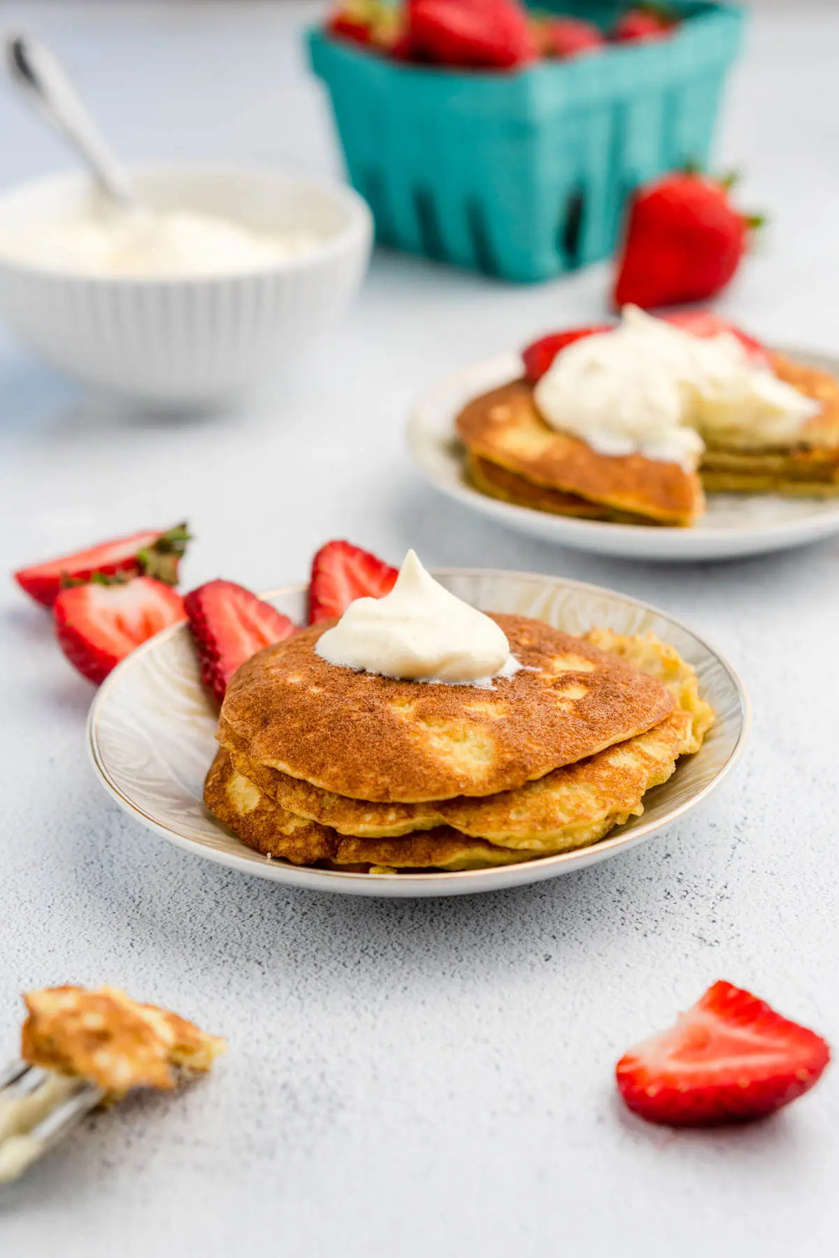 Pancakes topped with whipped cream and strawberries on plates near basket of fruit and bowl of whipped cream.